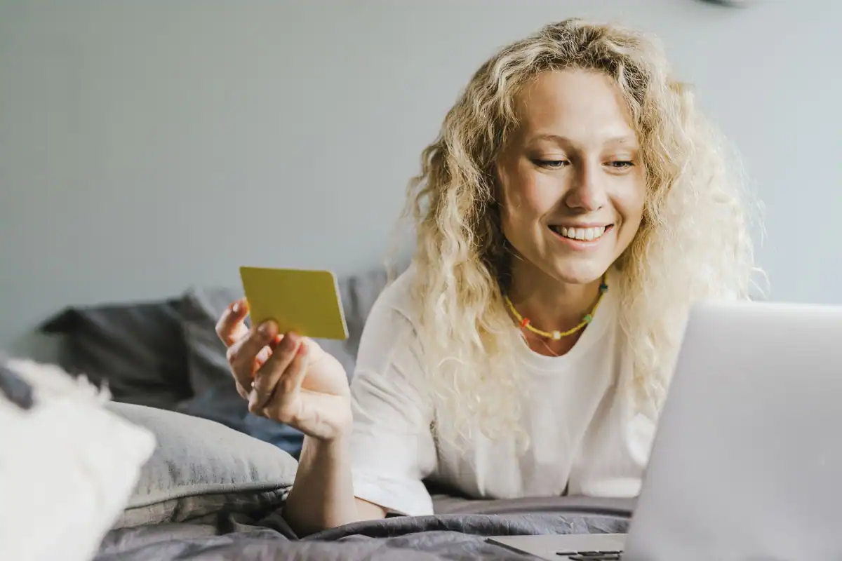 Femeie blondă și zâmbitoare face cumpărături online, cu un card bancar în mână